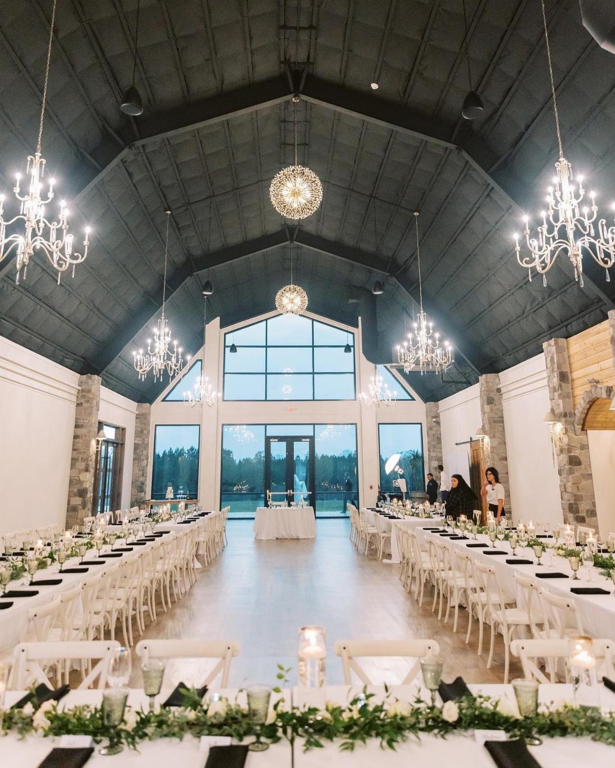 Long dining tables with white linens and greenery in a vaulted hall.