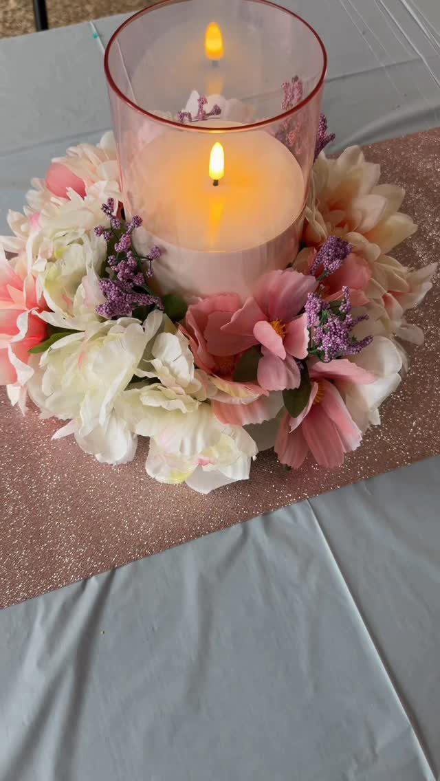 Lit pillar candle in glass cylinder surrounded by pink and white artificial flowers