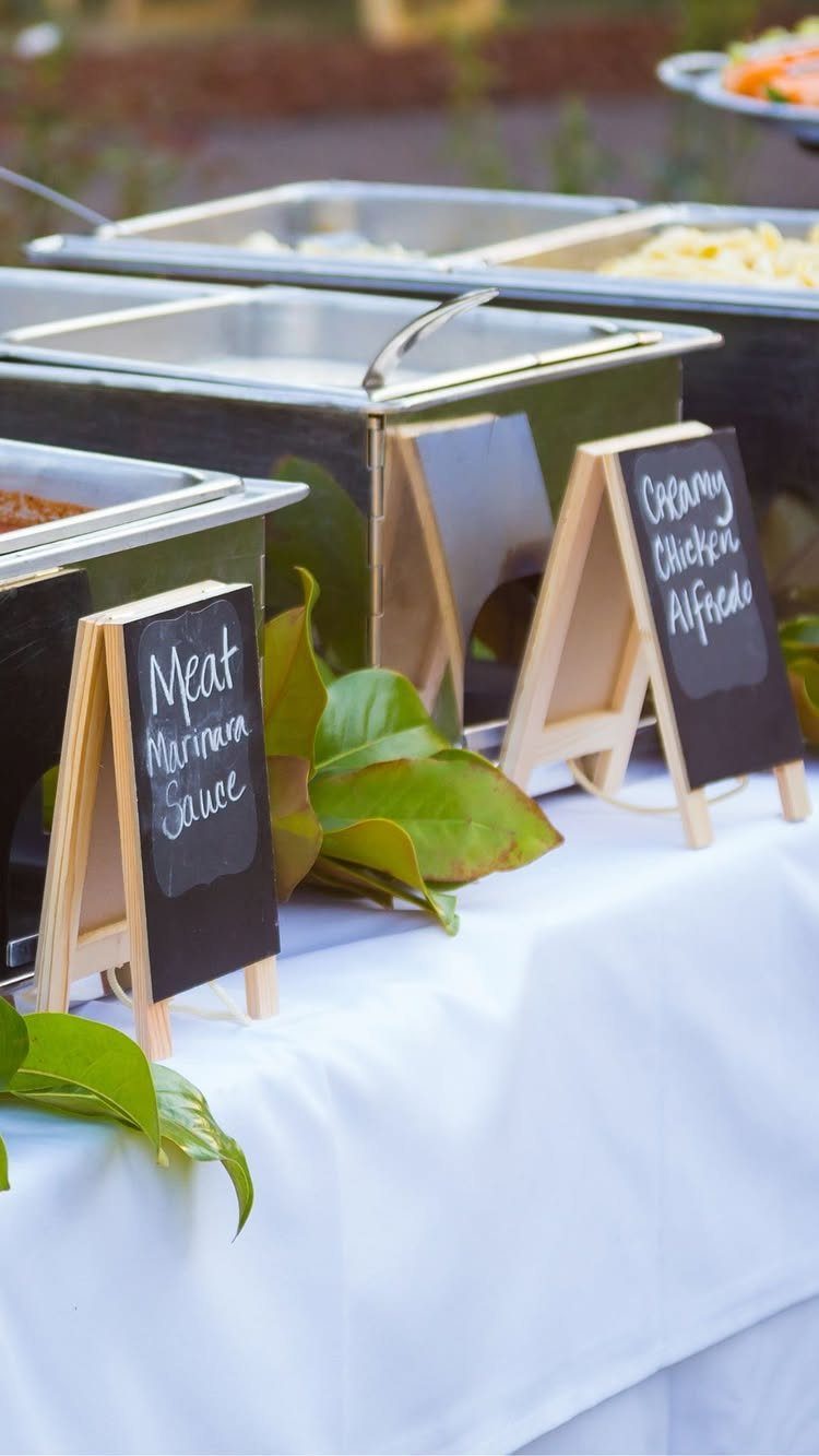 Small chalkboard signs on wooden easels labeling food in silver buffet chafing dishes.