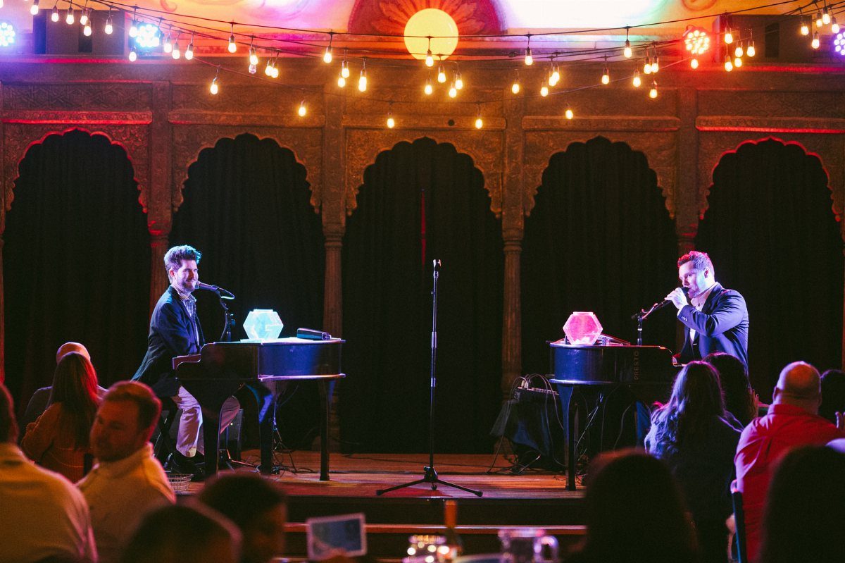Two musicians playing grand pianos and singing on a stage with string lights.