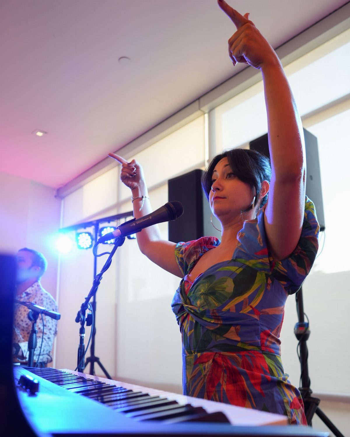 Woman in floral dress performing at a piano with stage lighting and microphone.
