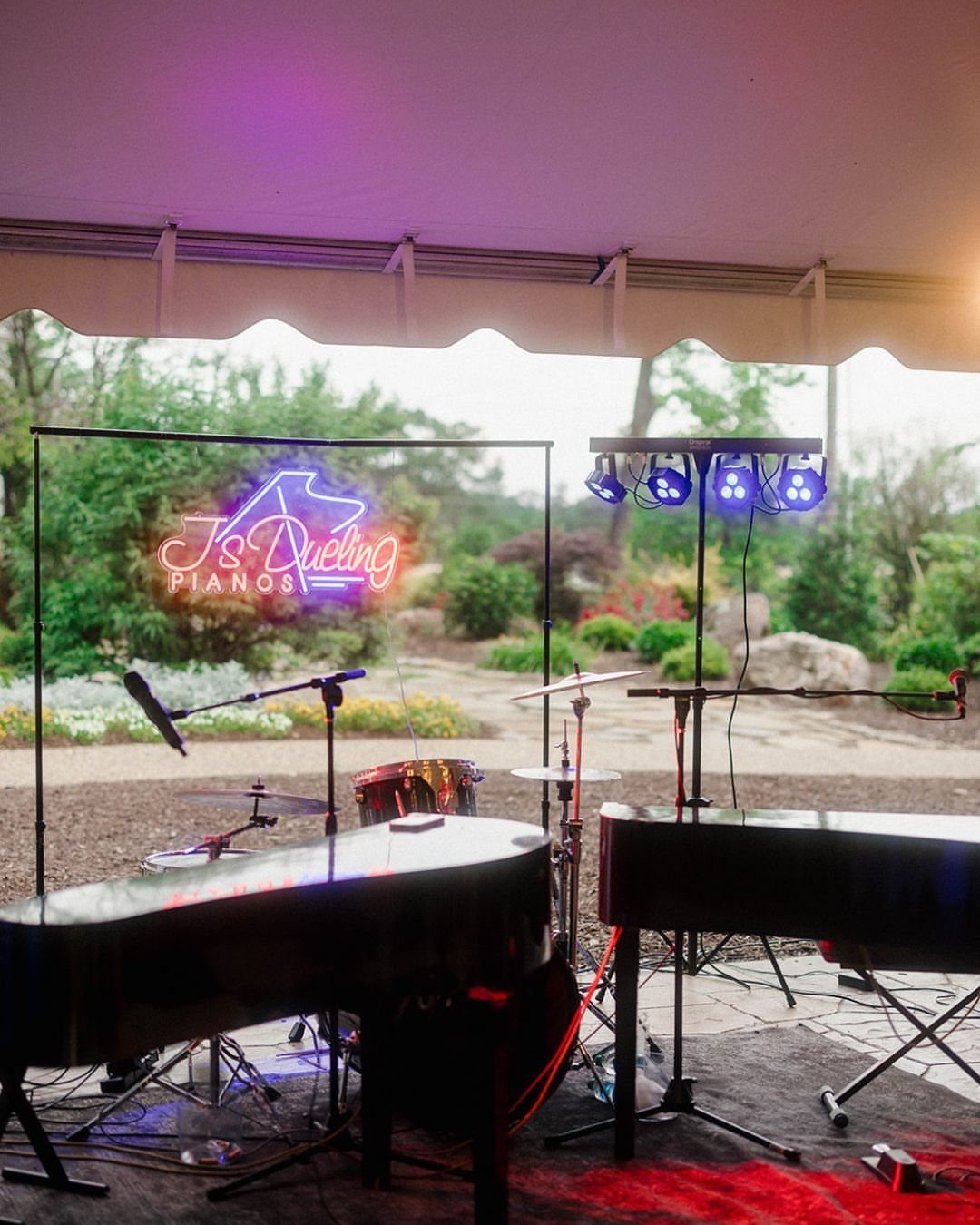 Two black pianos and a neon sign under a white outdoor event tent.