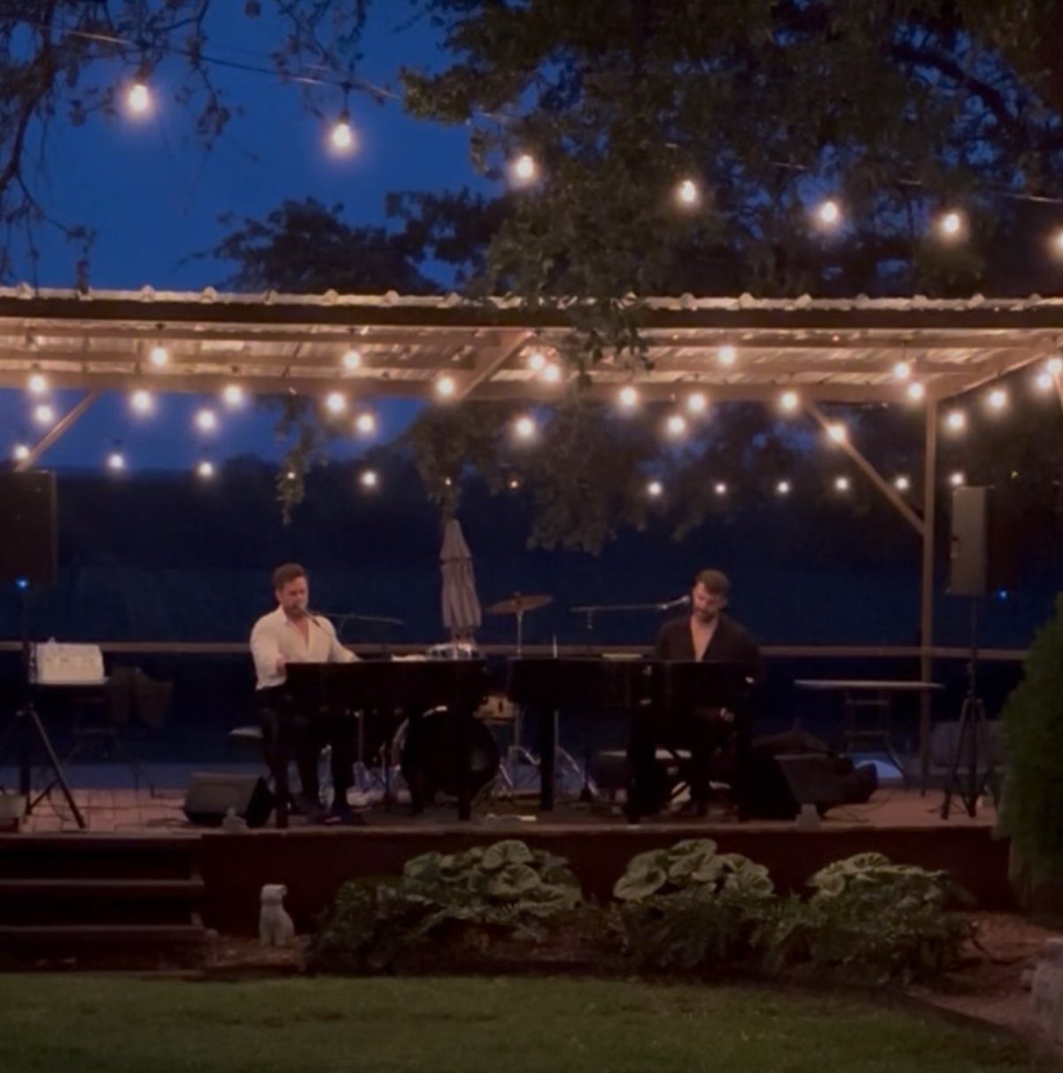 Two pianists performing on a wooden outdoor stage under warm string lights at dusk.
