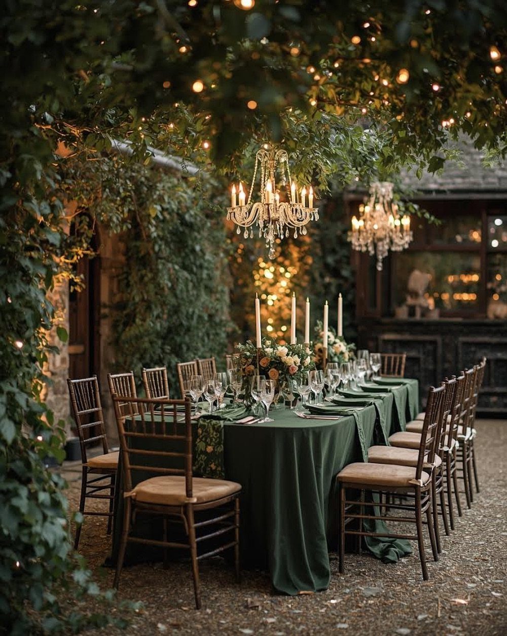 Long dining table with green cloth and candles under leafy trees and chandeliers.