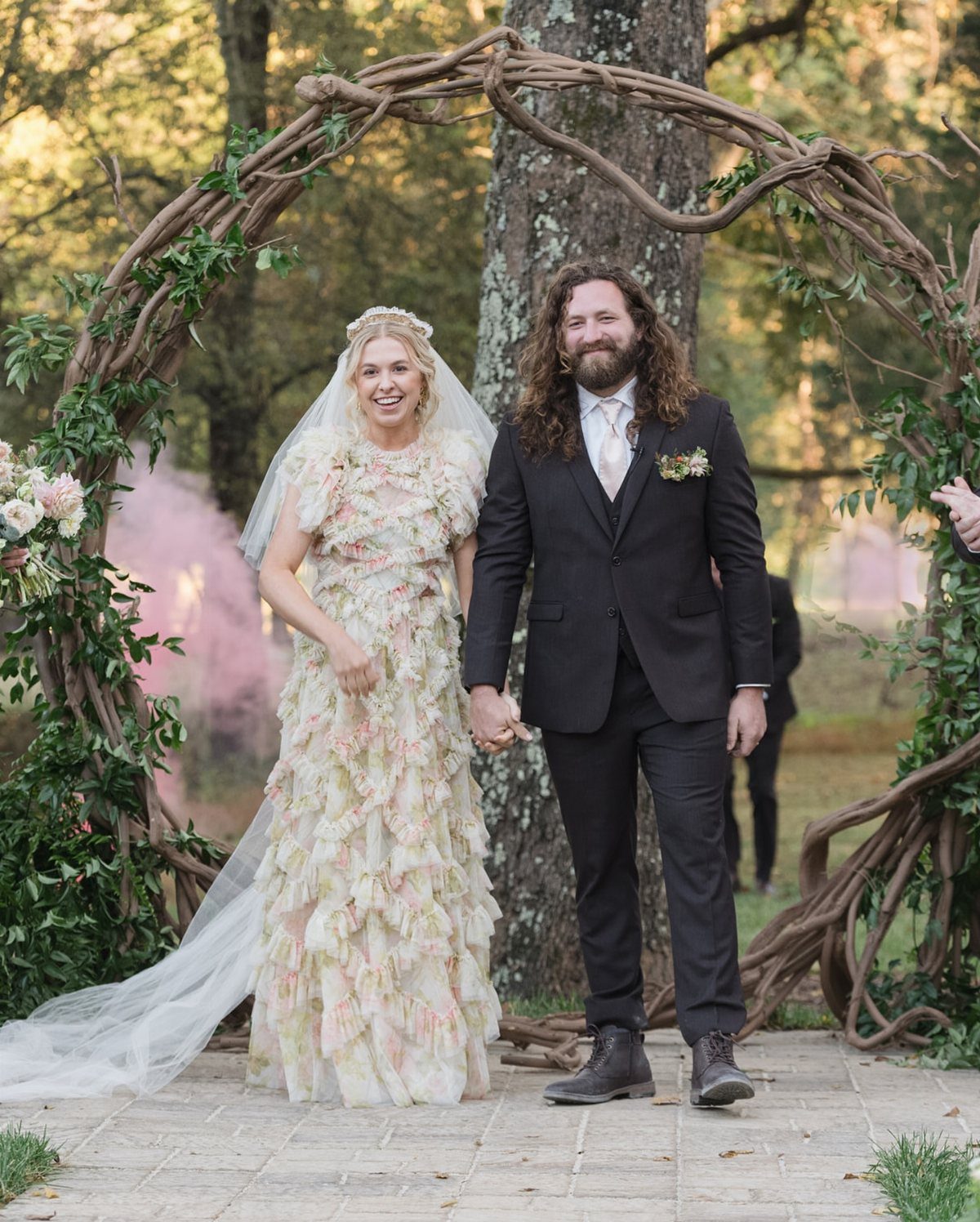 Bride in floral ruffled gown and groom in black suit under branch arch.