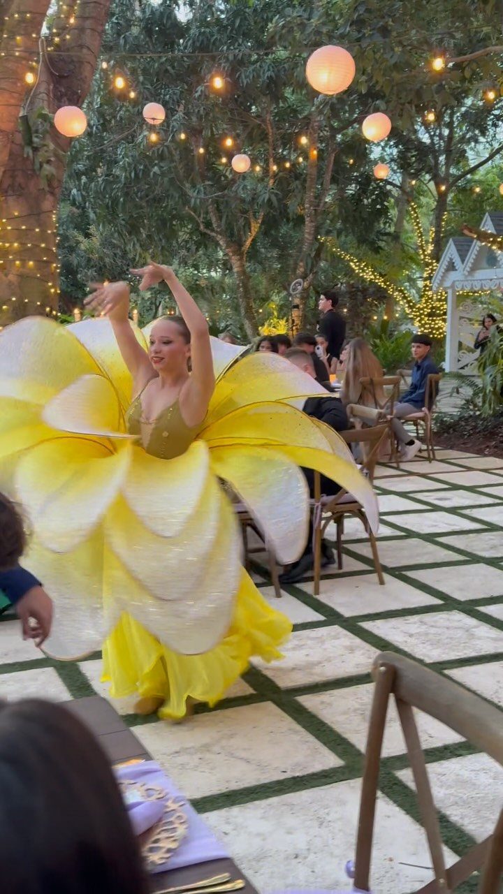 Woman in a yellow flower costume dancing at an outdoor garden event with guests.