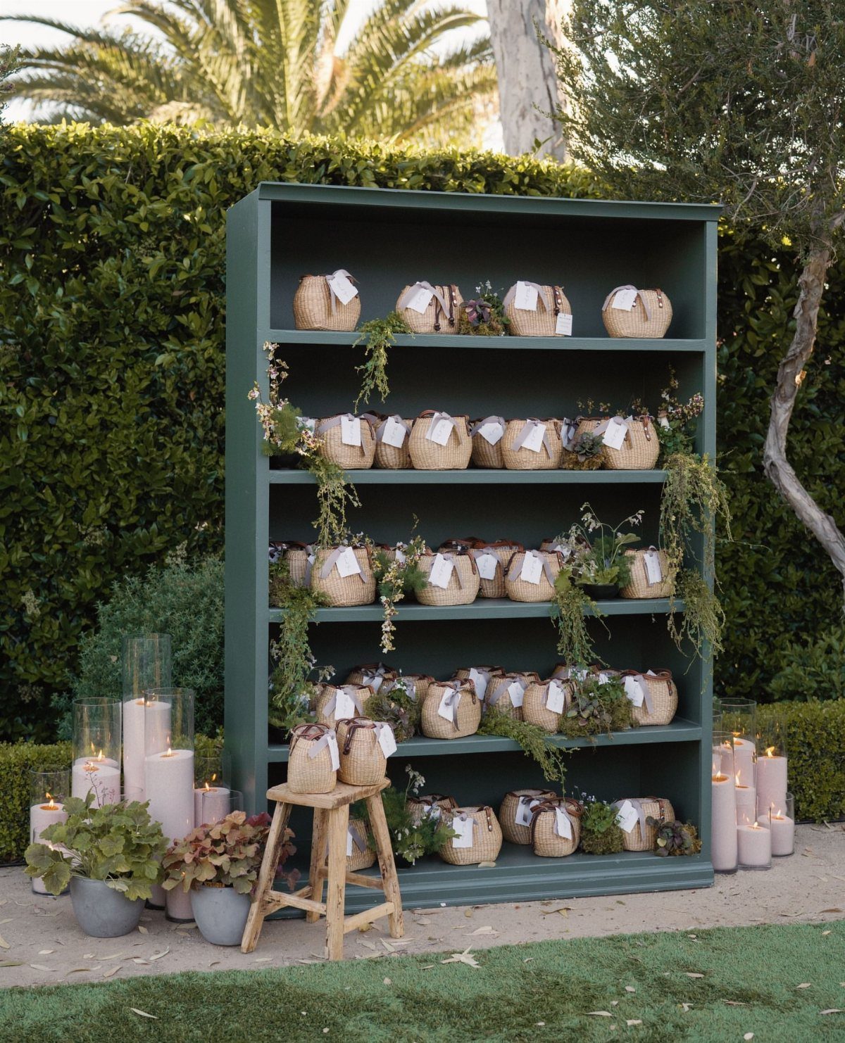 Burlap gift sacks with tags arranged on a green bookshelf outdoors with candles.