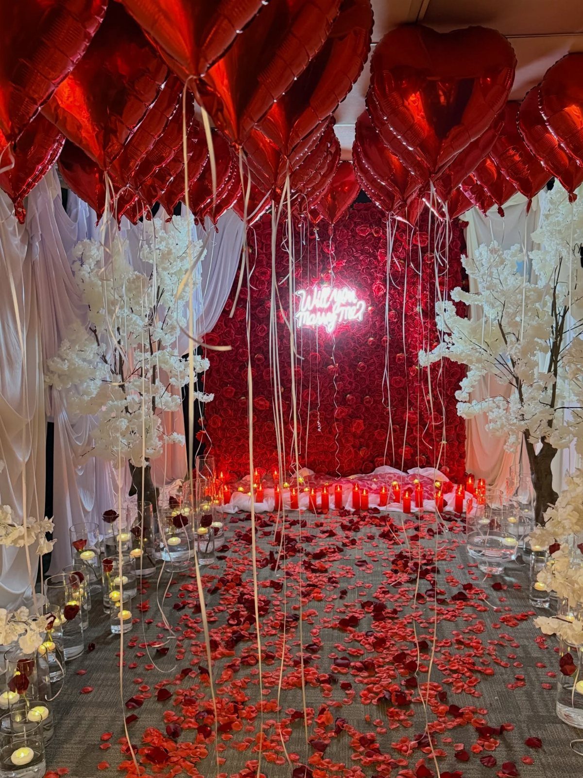 Romantic room decorated with red heart balloons, rose petals, and white blossom trees.