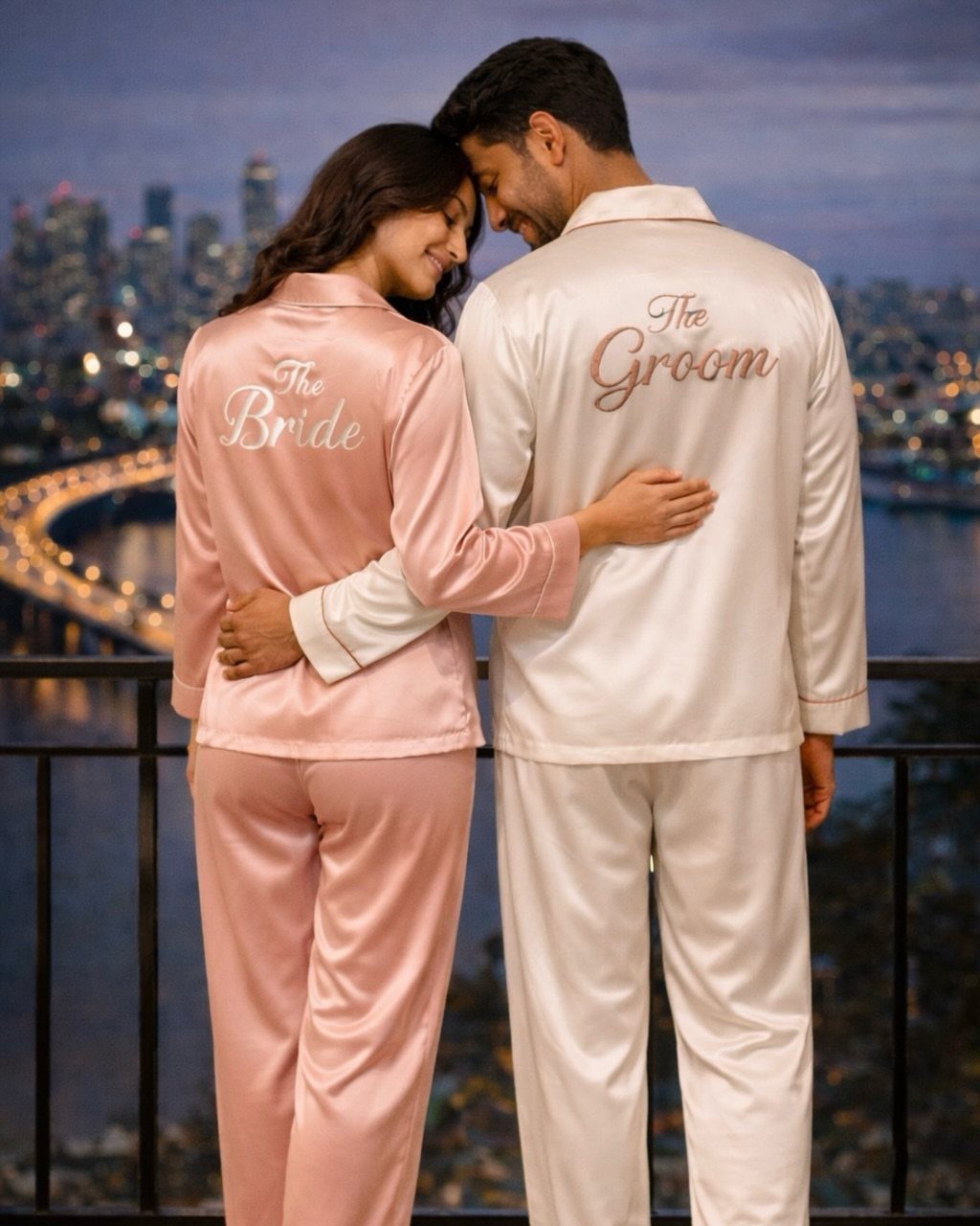 Couple in matching satin pajamas labeled The Bride and The Groom on a balcony.