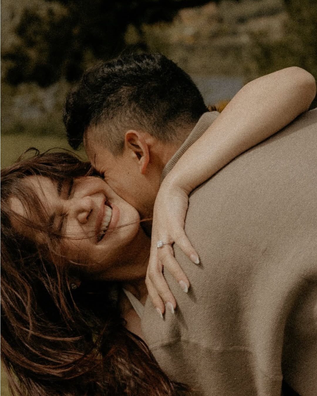 Smiling woman with engagement ring being embraced and kissed by her partner outdoors.