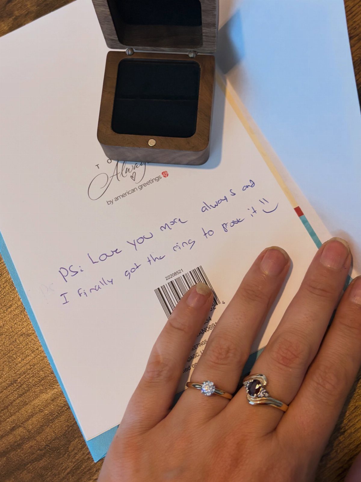 Hand wearing engagement rings resting on a handwritten love note and ring box.
