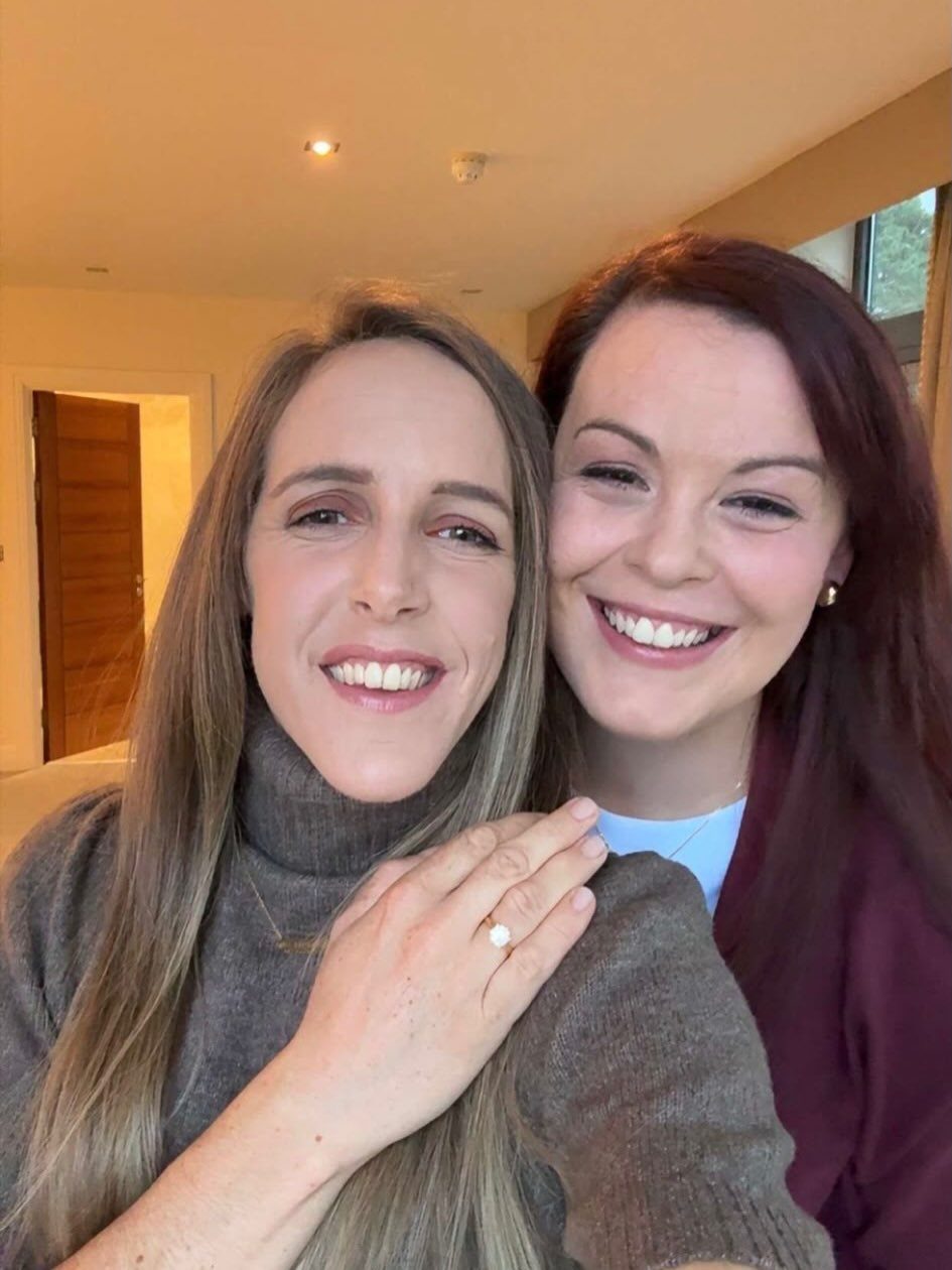 Two smiling women posing together with a diamond engagement ring visible on a hand.