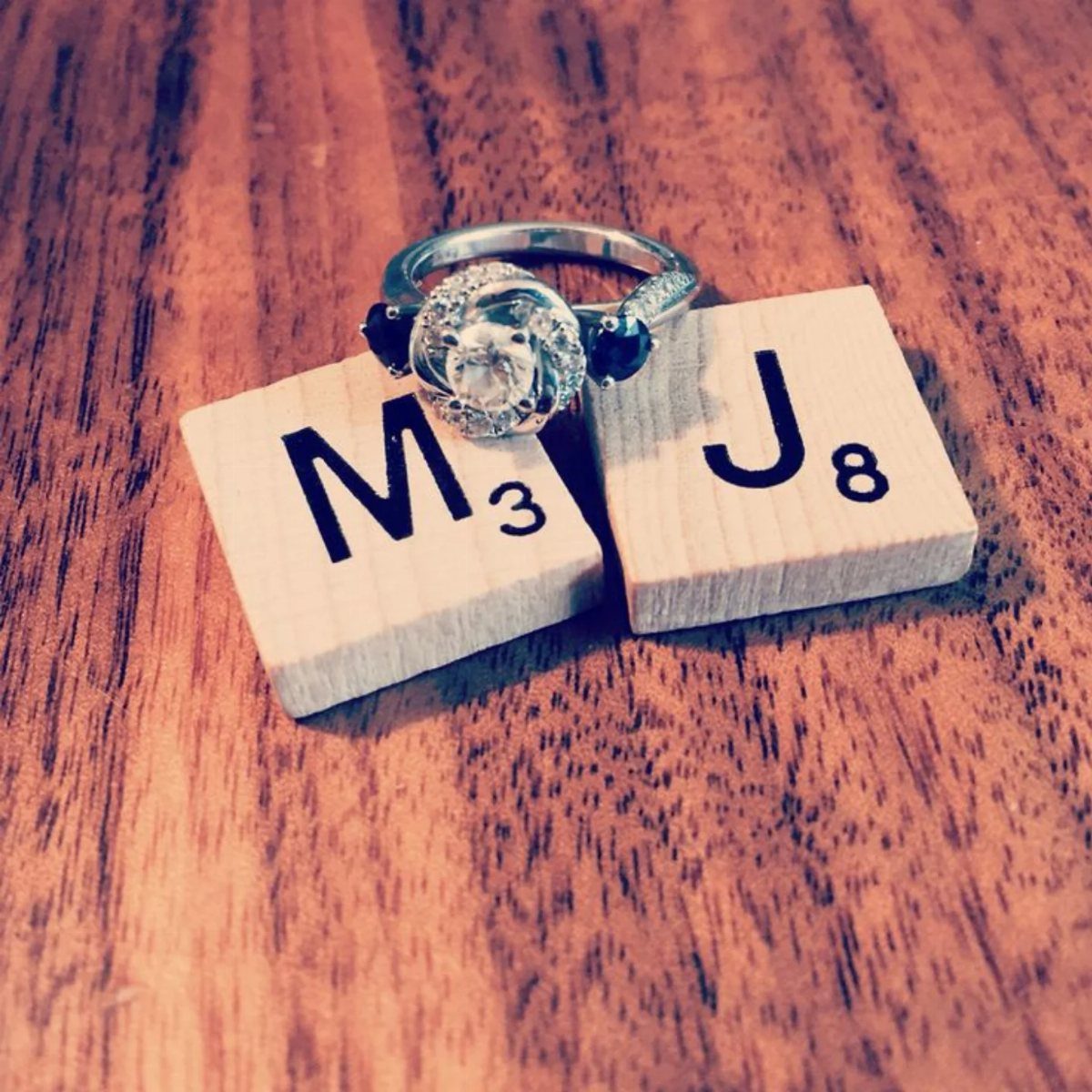 Engagement ring resting on Scrabble tiles with letters M and J on wood.
