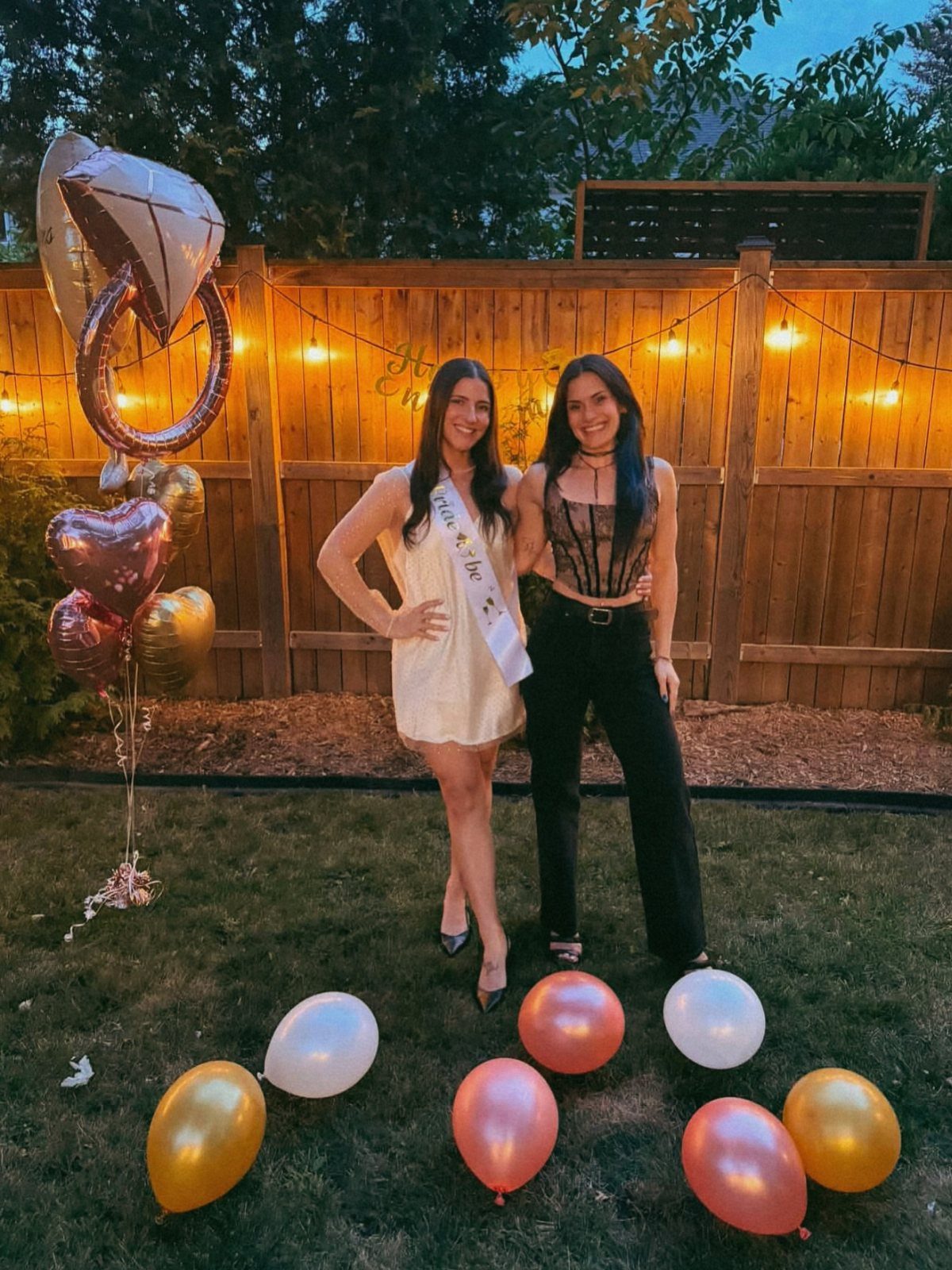 Two women posing by a large diamond ring balloon and string lights outdoors.