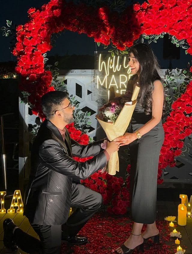 Man kneeling to propose to a woman inside a red rose heart arch.