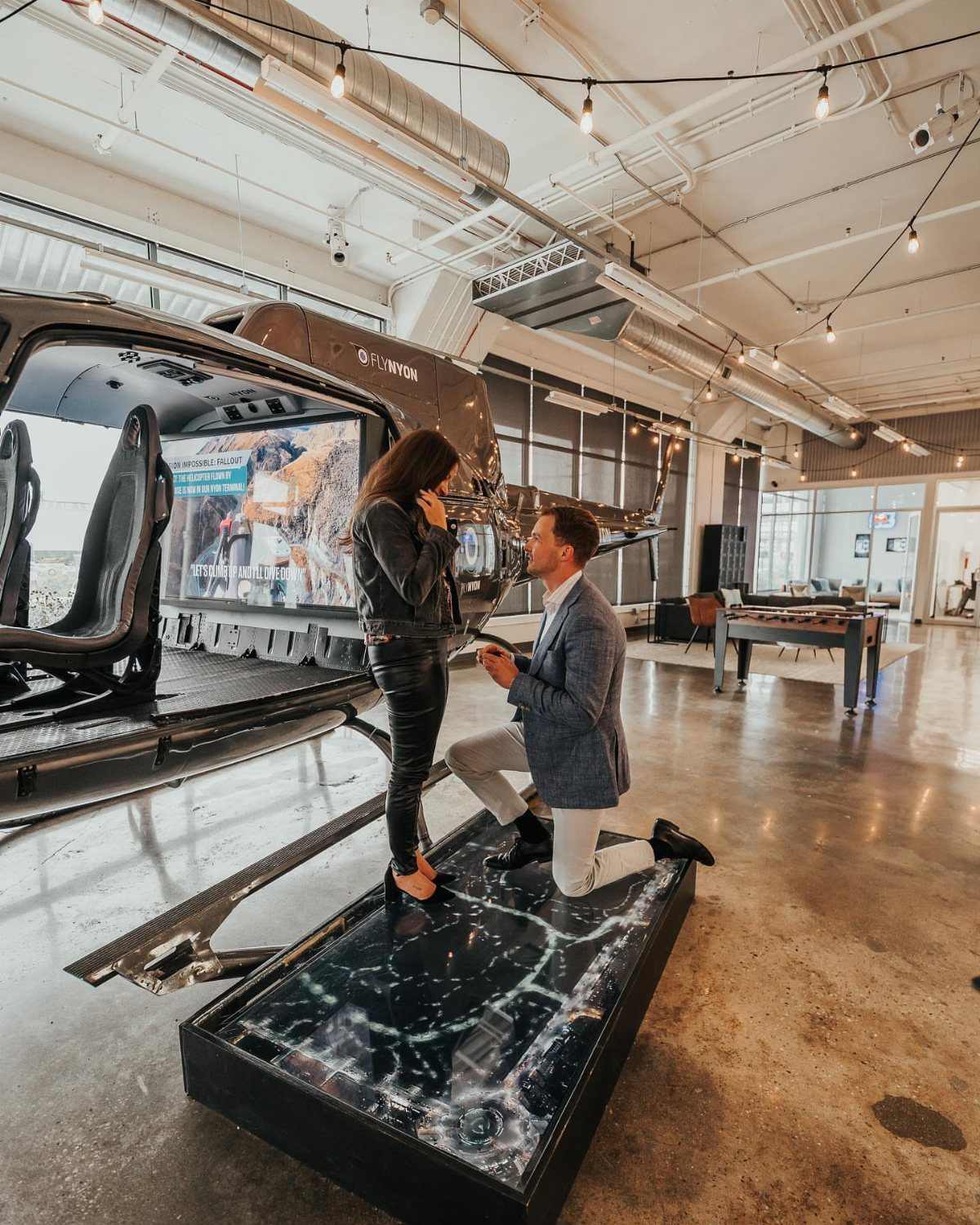Man kneeling to propose to a woman inside a hangar with a helicopter.