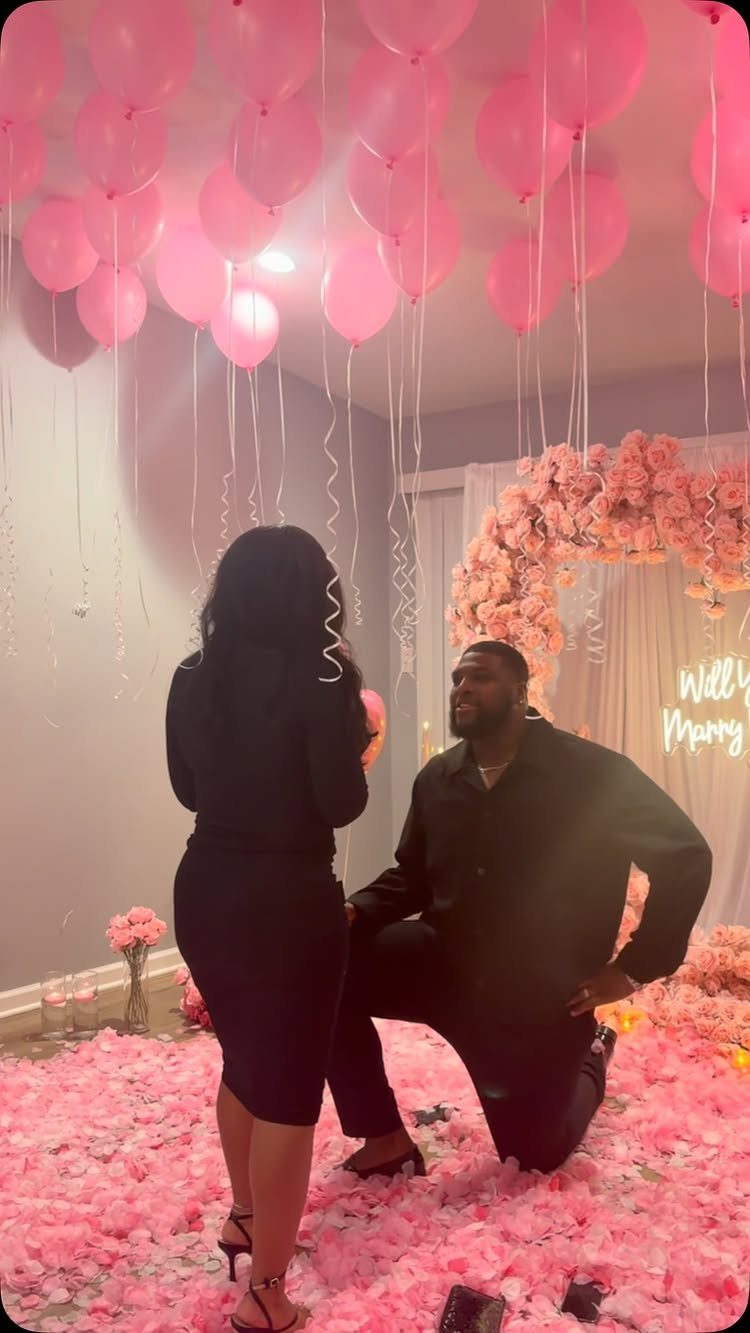 Man kneeling on pink petals proposing to a woman under floating pink balloons.