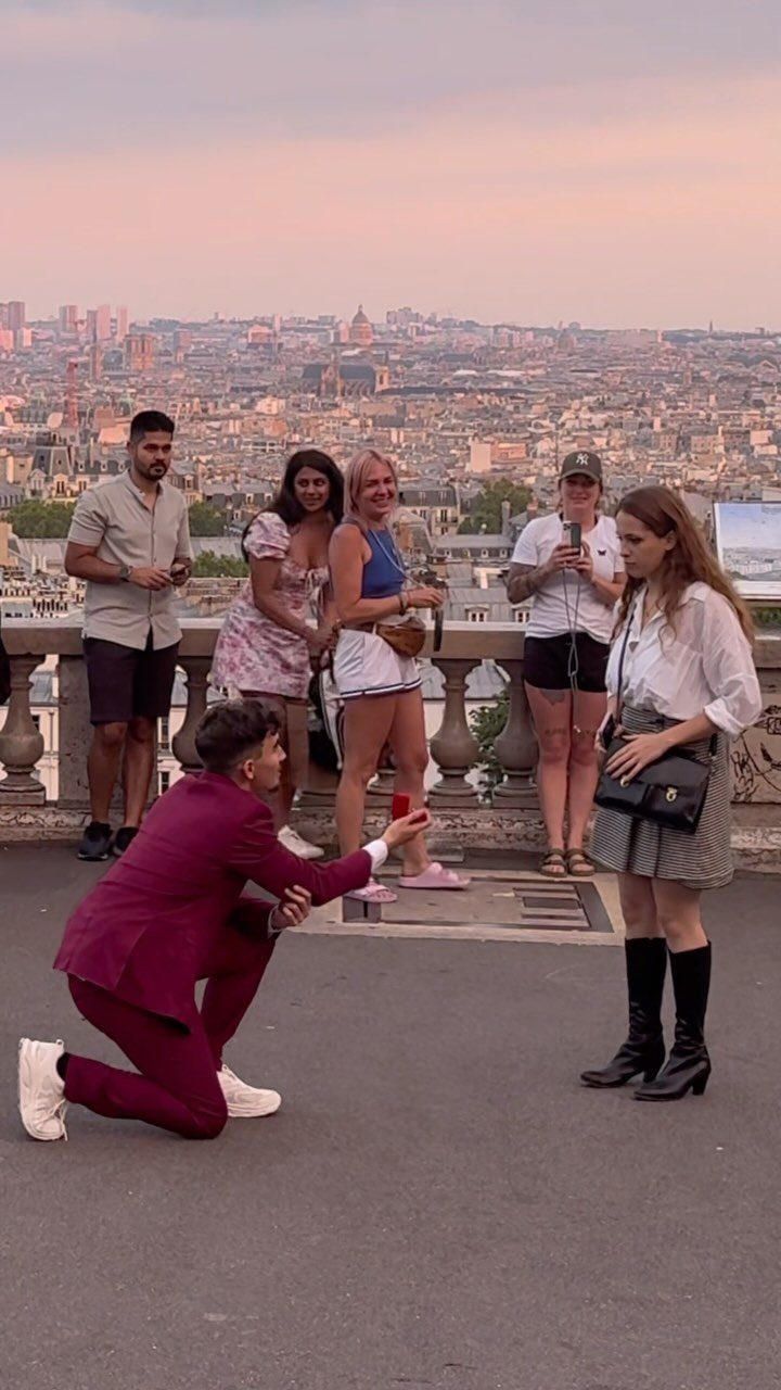 Man in maroon suit proposing to woman on a balcony overlooking Paris skyline