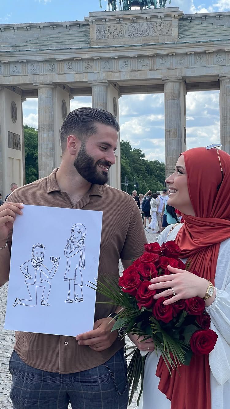 Couple holding a proposal drawing and red roses before the Brandenburg Gate.