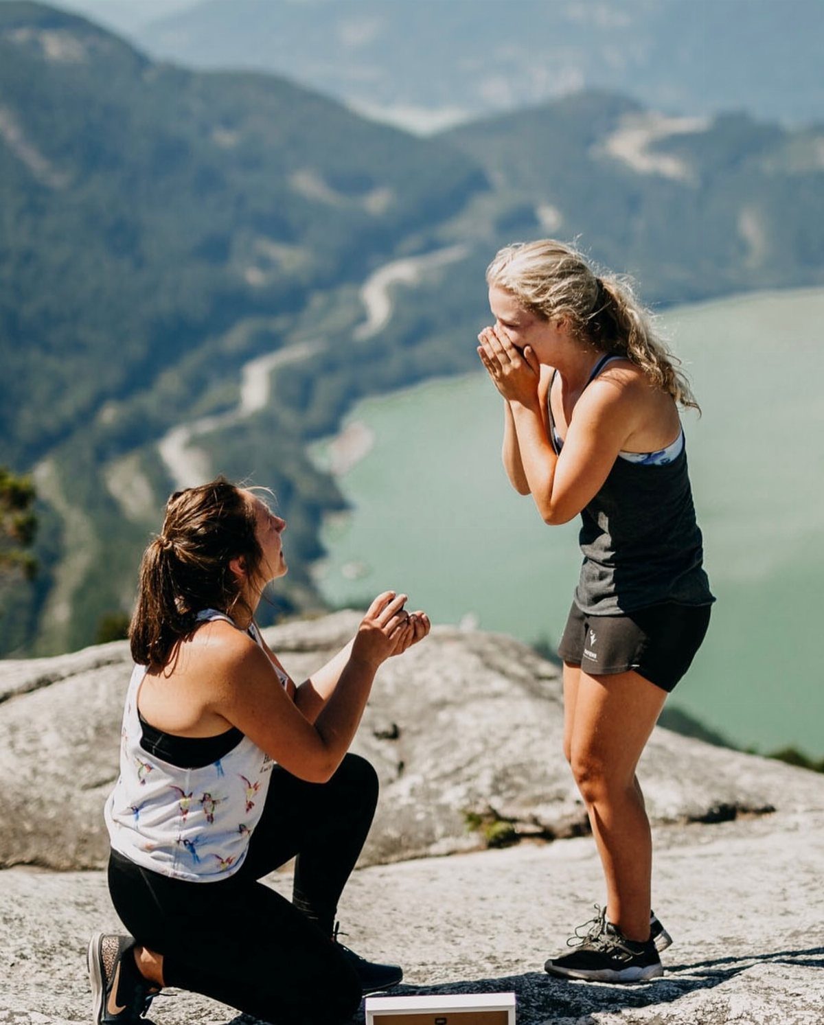 Woman kneeling to propose to her partner on a rocky mountain overlooking a lake.