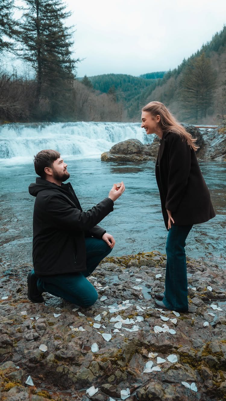 Man kneeling on rocky riverbank proposing to a woman with a waterfall background