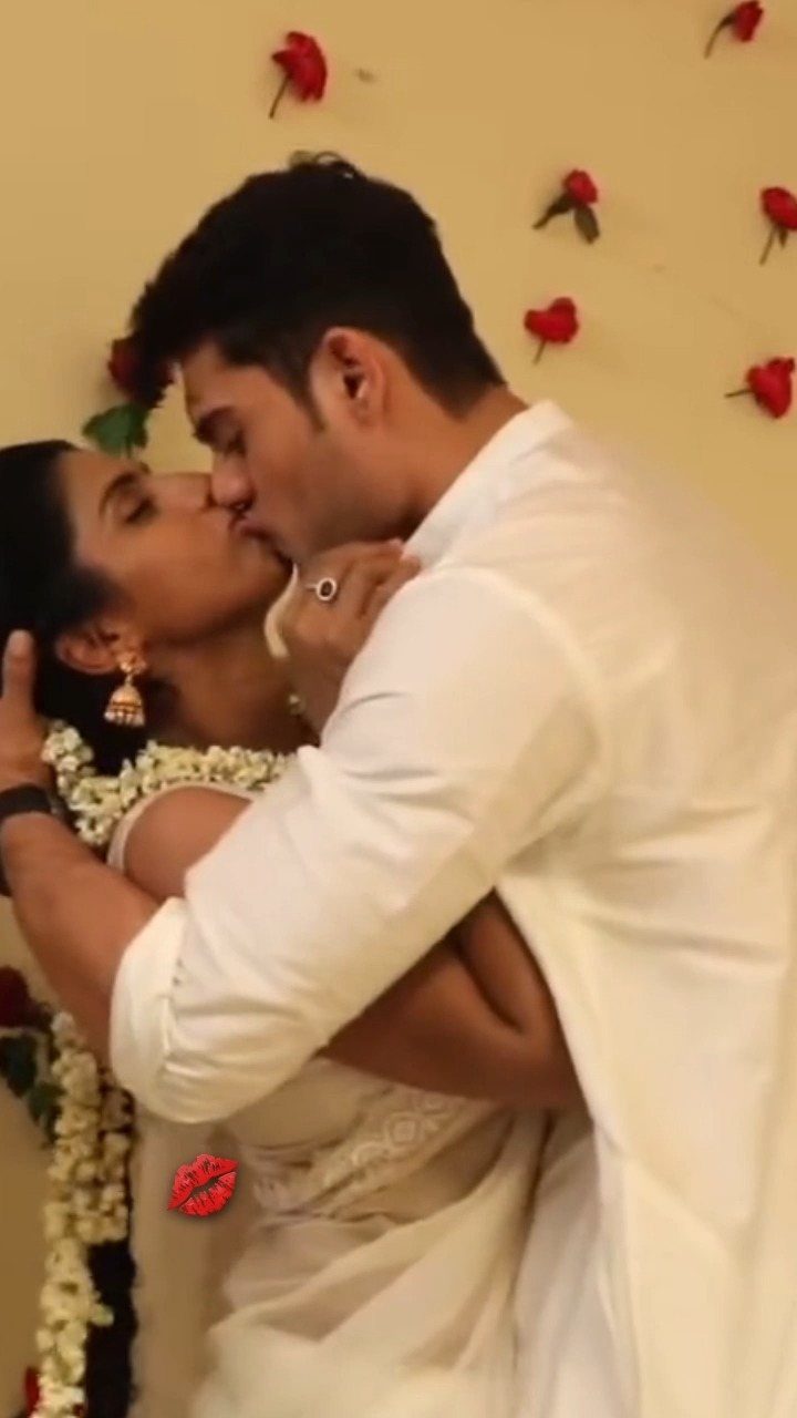 Couple in white wedding attire kissing with their eyes closed against rose wall