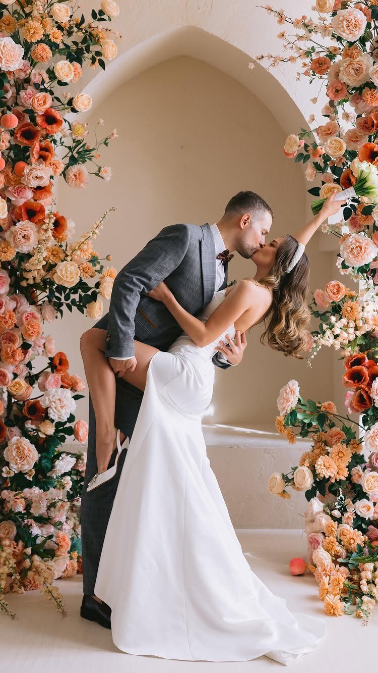Groom dipping and kissing bride under a peach and orange floral arch.