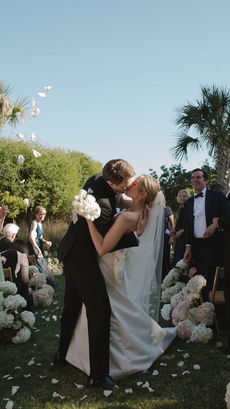 Bride and groom kissing outdoors on grass with white petals falling around them.