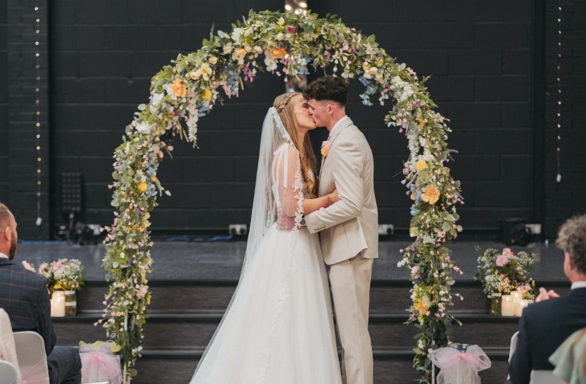 Bride and groom kissing under a floral arch during their wedding ceremony.