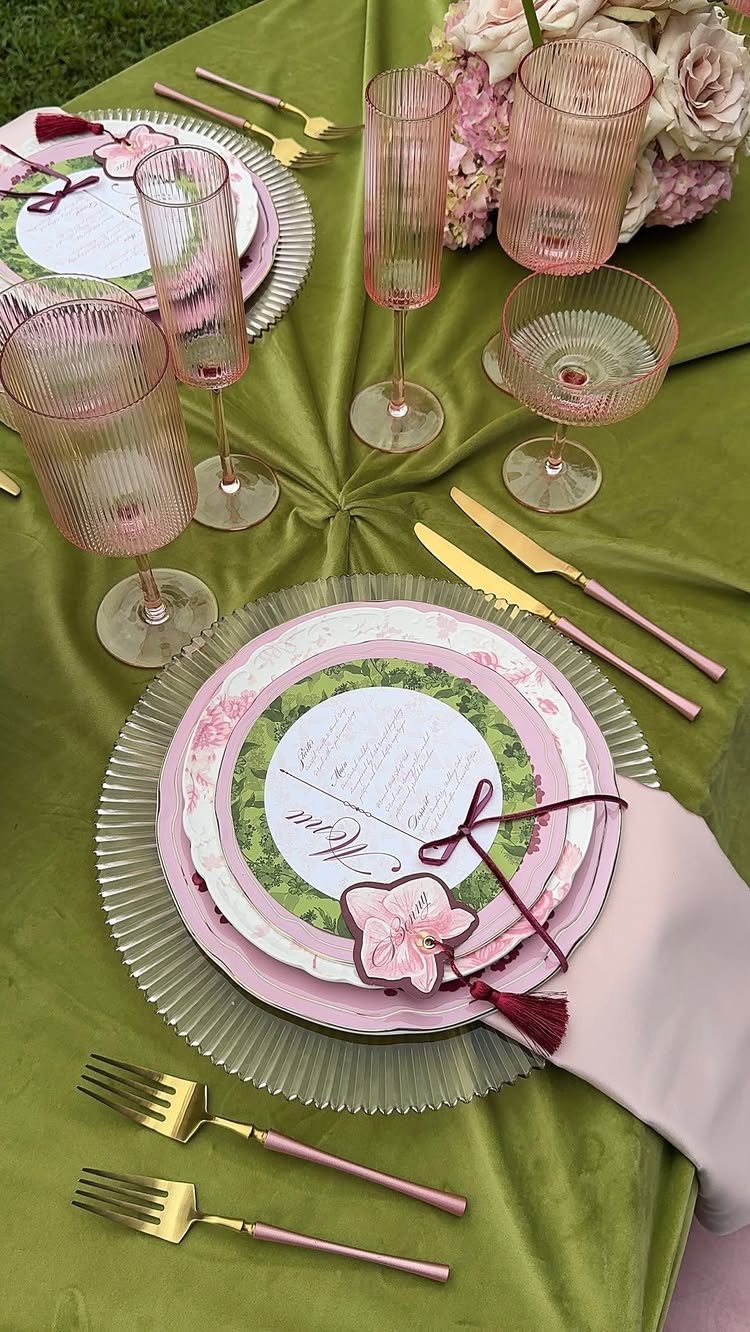 Formal outdoor table setting with green satin cloth, pink glassware, and gold cutlery.