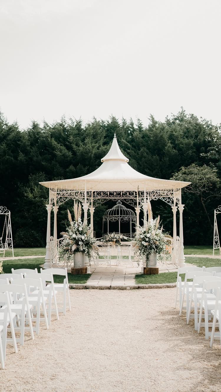 White ornate wedding gazebo with floral arrangements and rows of white folding chairs.
