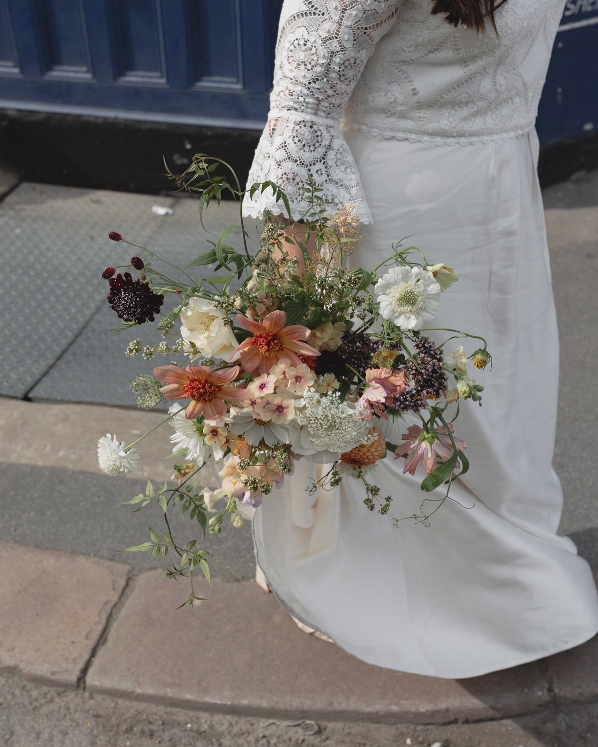 Loose organic wedding bouquet with peach and burgundy flowers held by a bride.