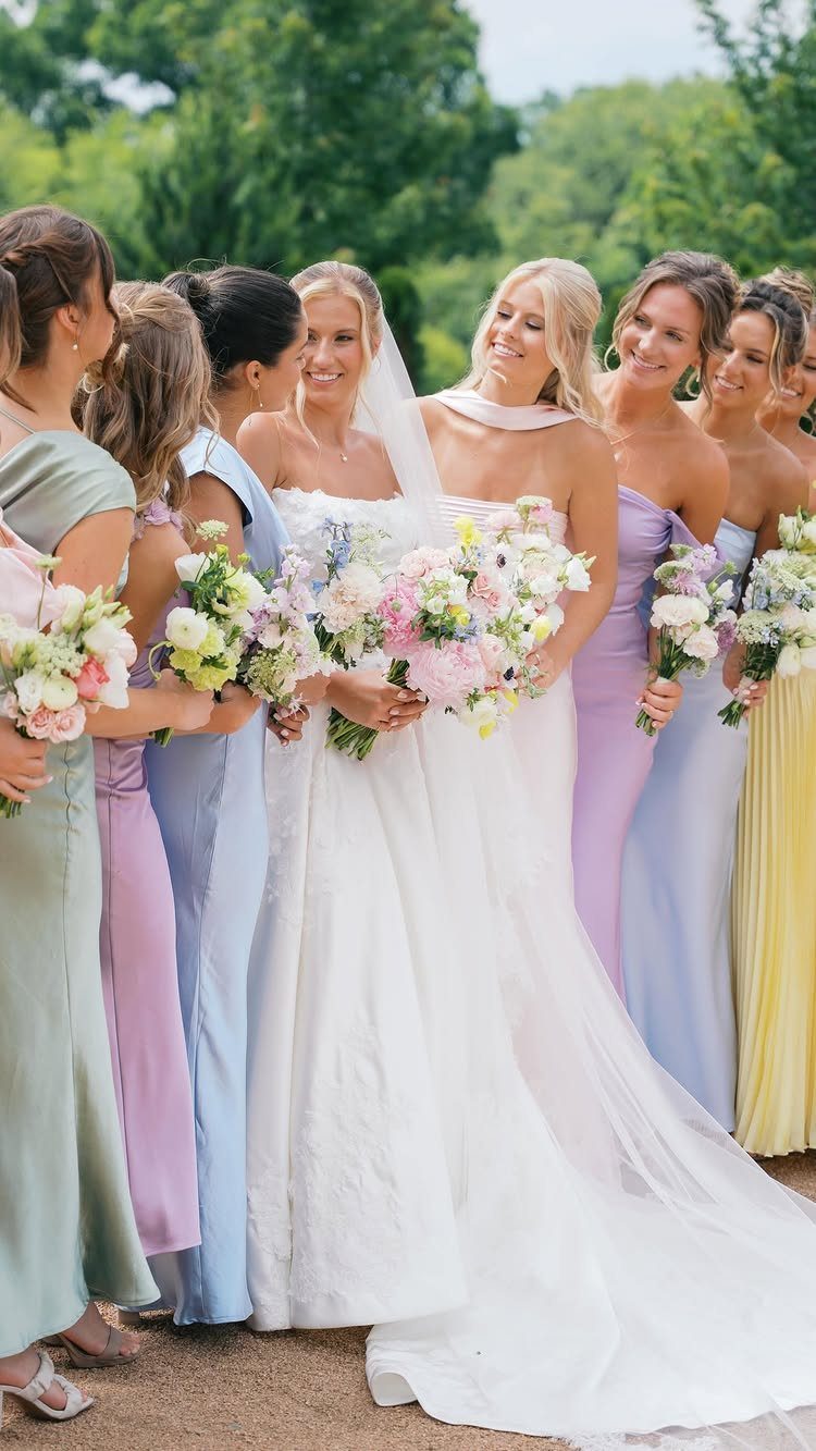 Bride and bridesmaids in mismatched pastel dresses holding bouquets in a green garden