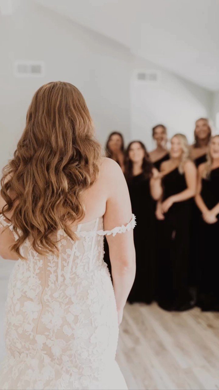 Bride in white lace dress facing bridesmaids wearing black dresses in a room.