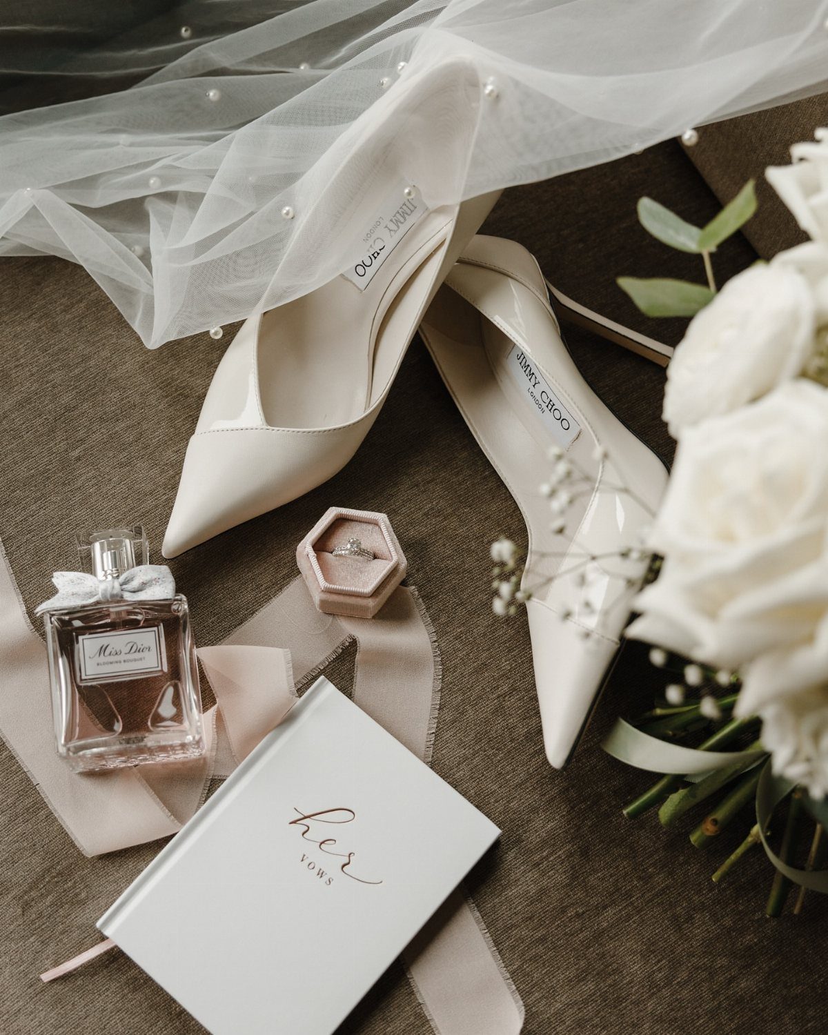 Wedding shoes, perfume, a ring, and a vow book arranged with a white veil.