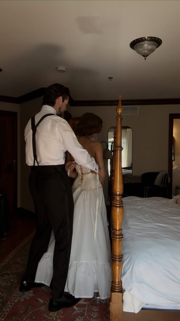 Groom helping bride with her wedding dress in a bedroom setting.