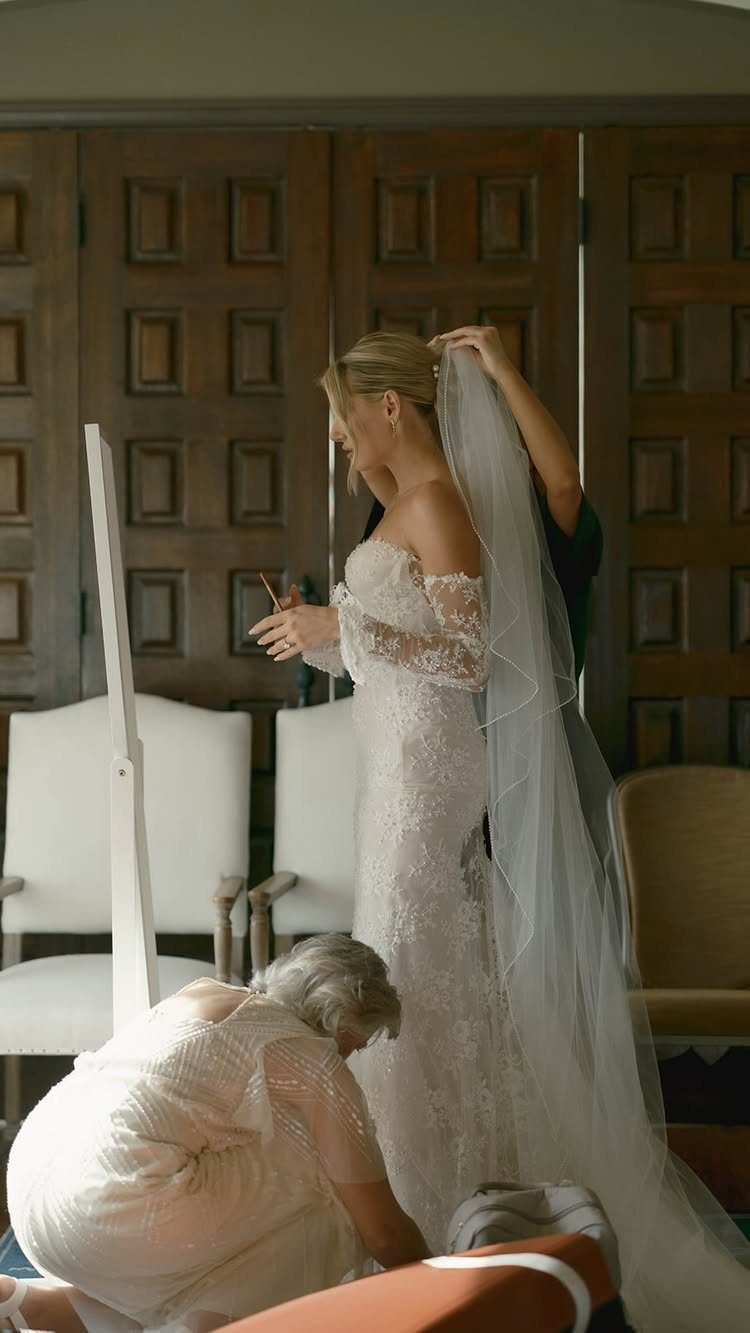Bride in lace wedding dress being helped with her veil and gown by others.
