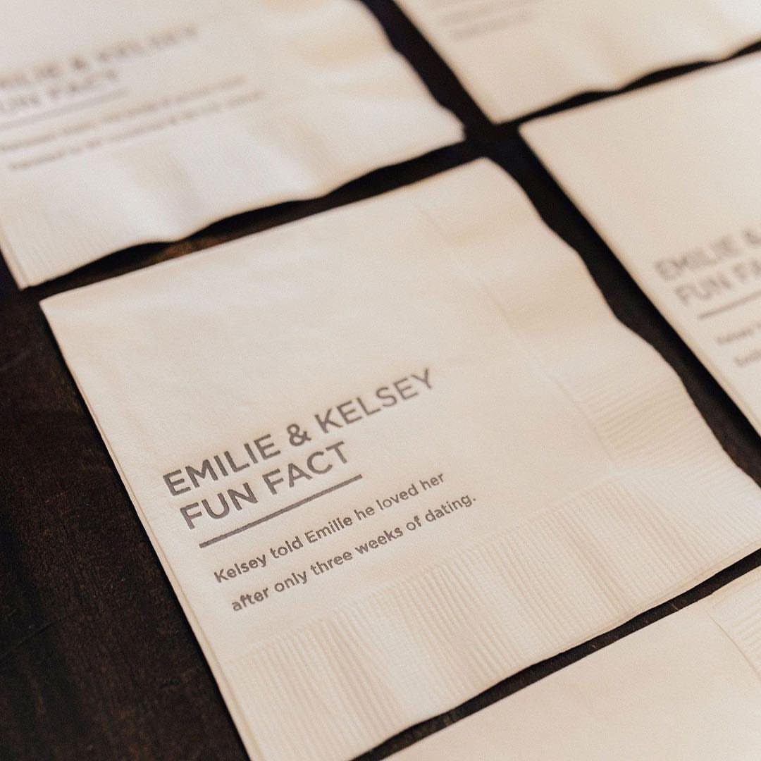White cocktail napkins with printed fun facts about a couple on dark wood.