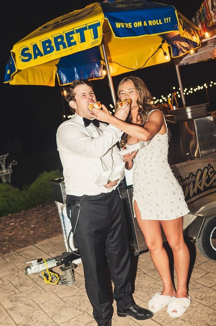 Bride and groom eating hot dogs from a food cart at their wedding reception.