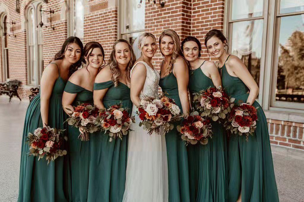 Bride in white gown with bridesmaids in emerald green dresses holding red bouquets.