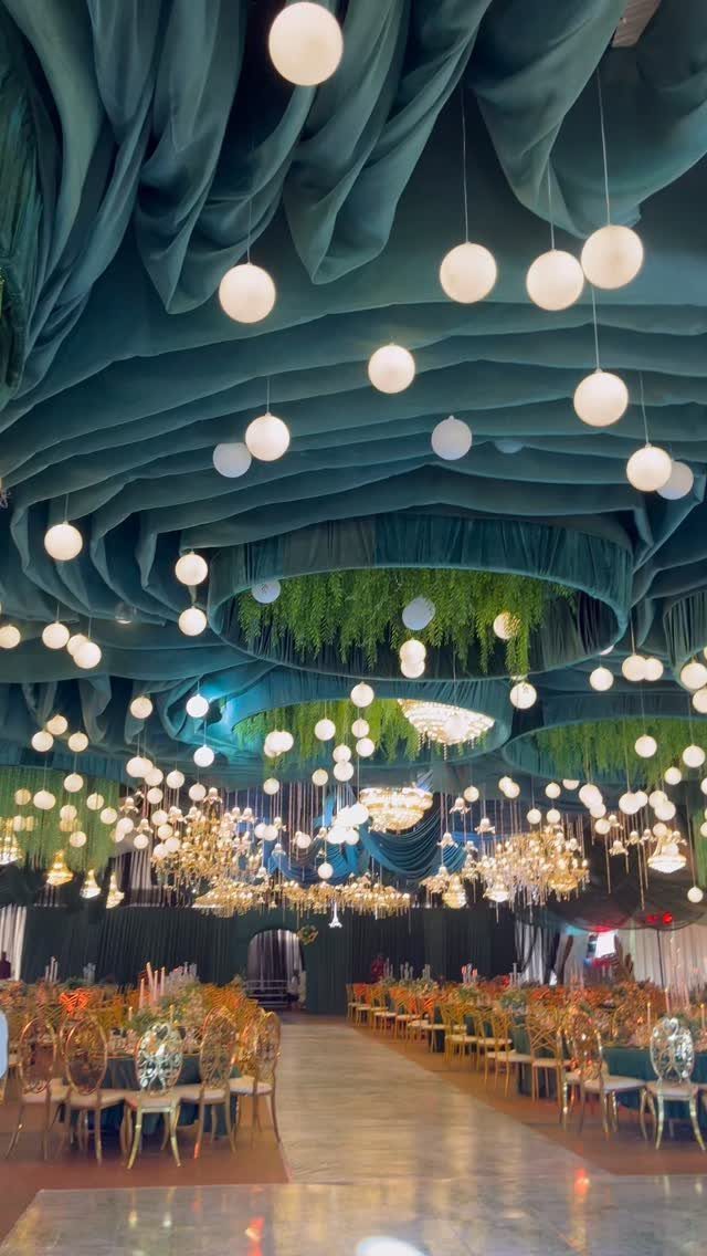 Deep teal ceiling drapes and hanging globe lights over gold wedding reception chairs