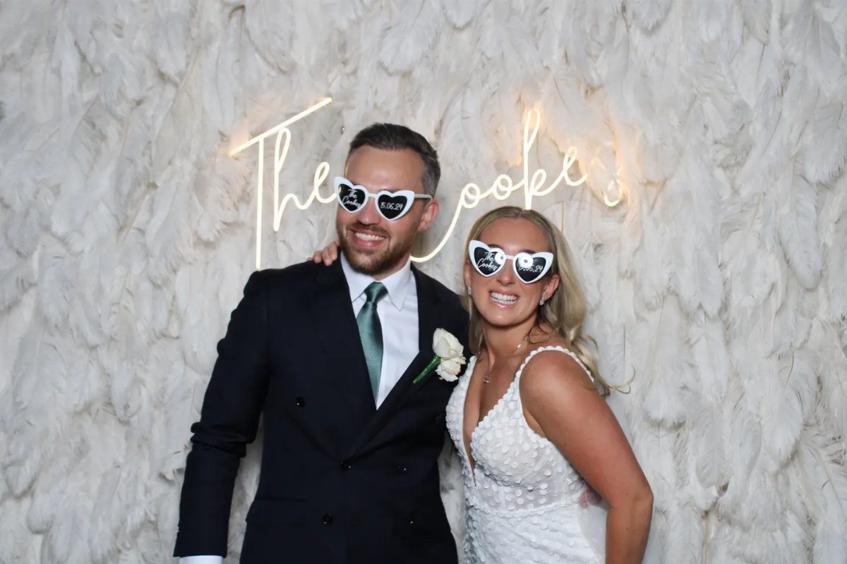 Bride and groom wearing heart sunglasses posing before a white feather photo backdrop