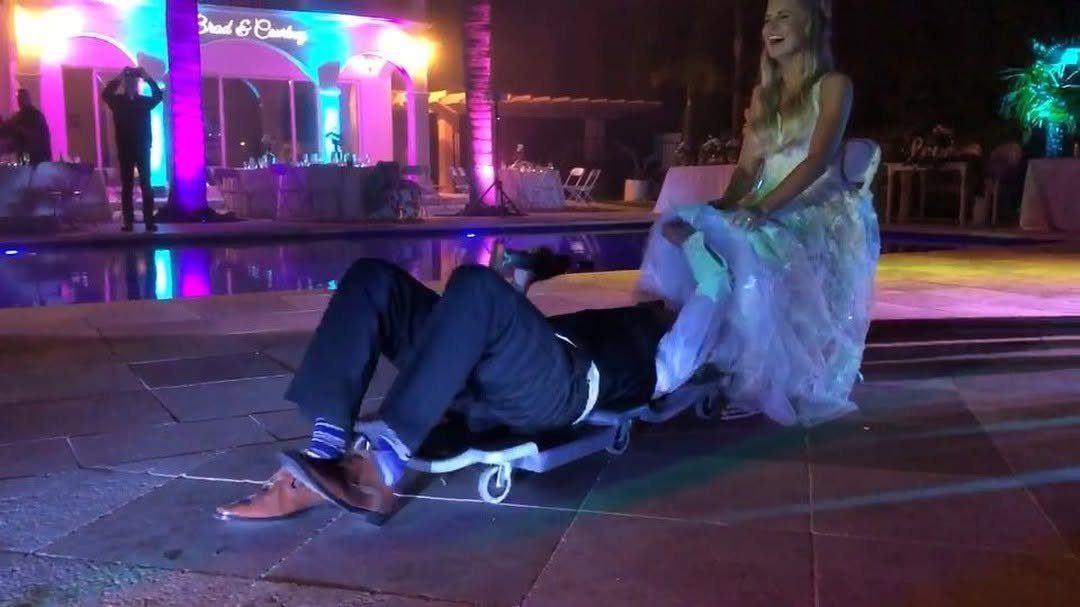 Bride laughing as groom slides toward her on a wheeled dolly at wedding.