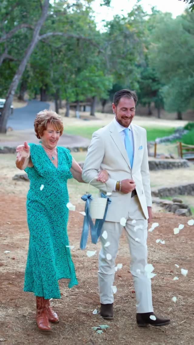 Older woman in green dress tossing flower petals while walking with a groom.
