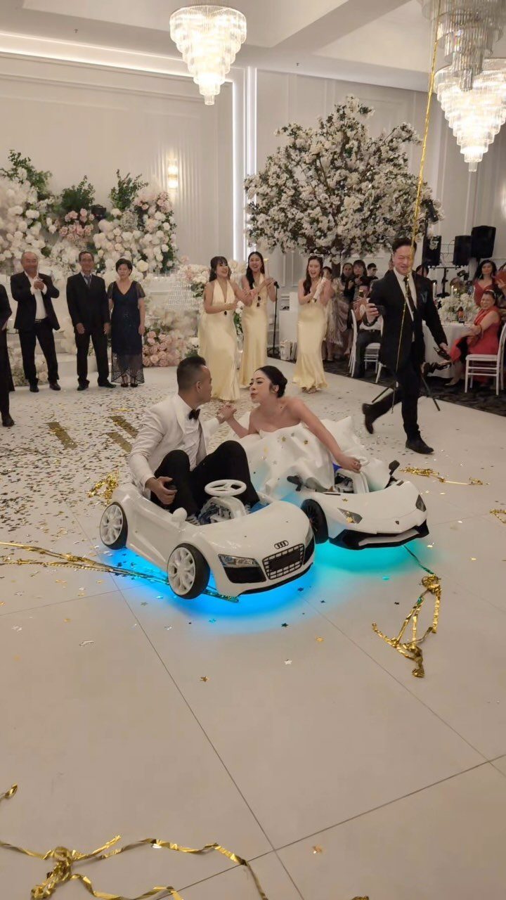 Bride and groom sitting in small white toy cars on a wedding dance floor