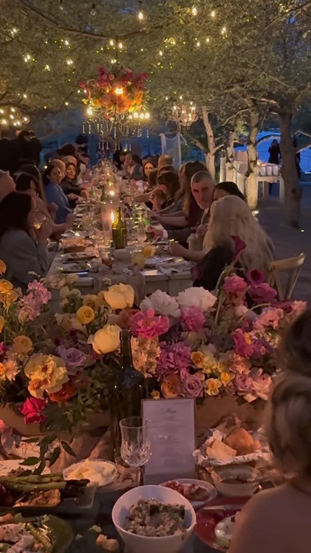 Outdoor dinner party with long tables, colorful flowers, and string lights under trees.