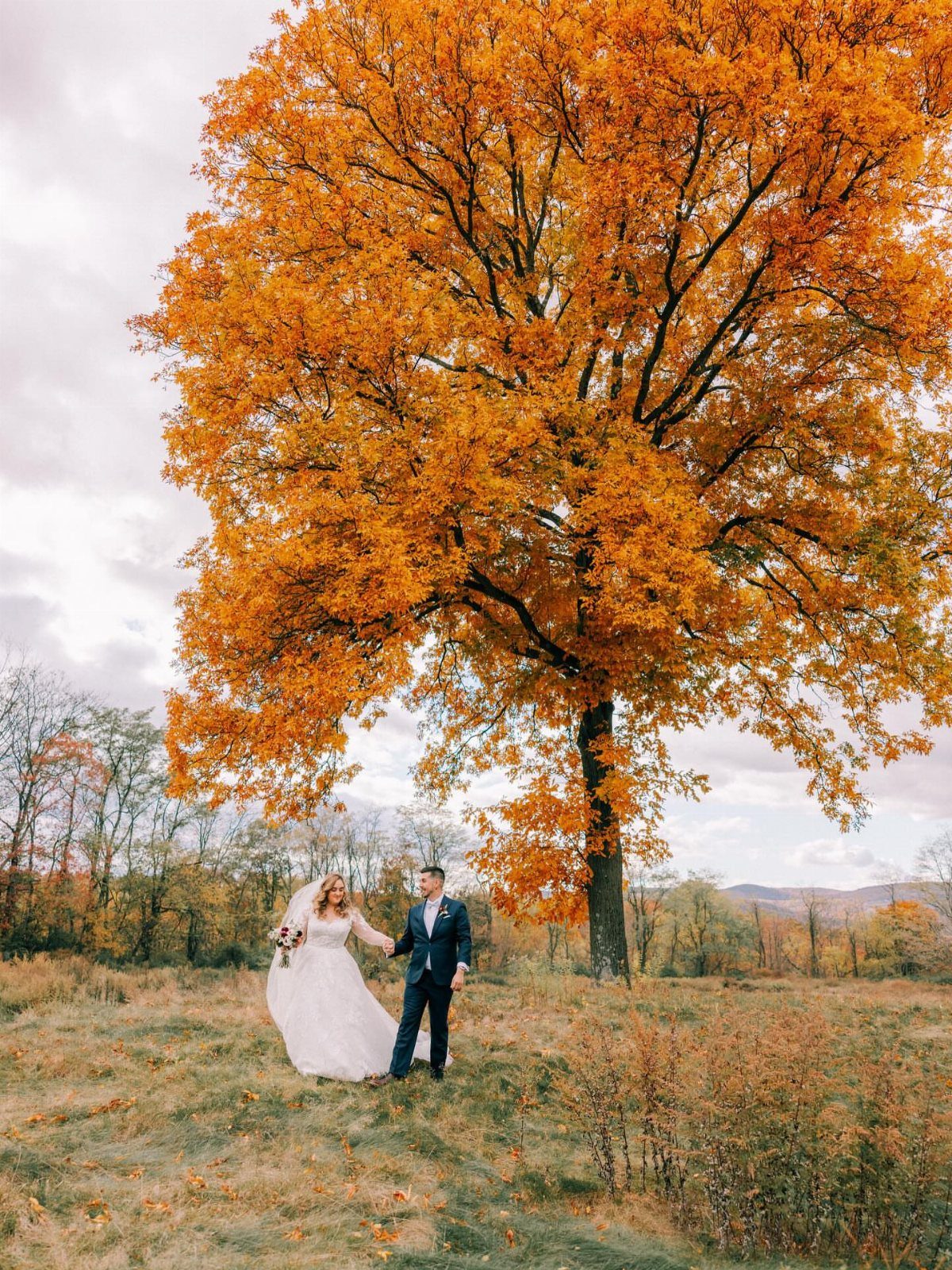 Bride and groom holding hands under a large orange autumn tree in a field.