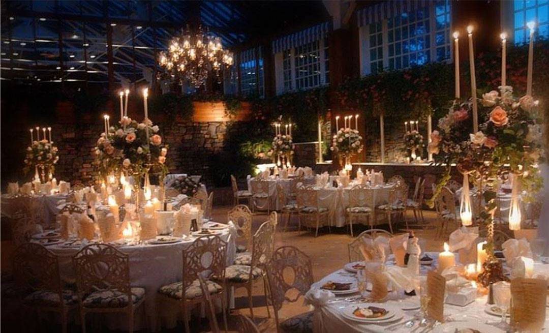 Wedding reception tables with white linens, floral centerpieces, and candles under a glass ceiling.