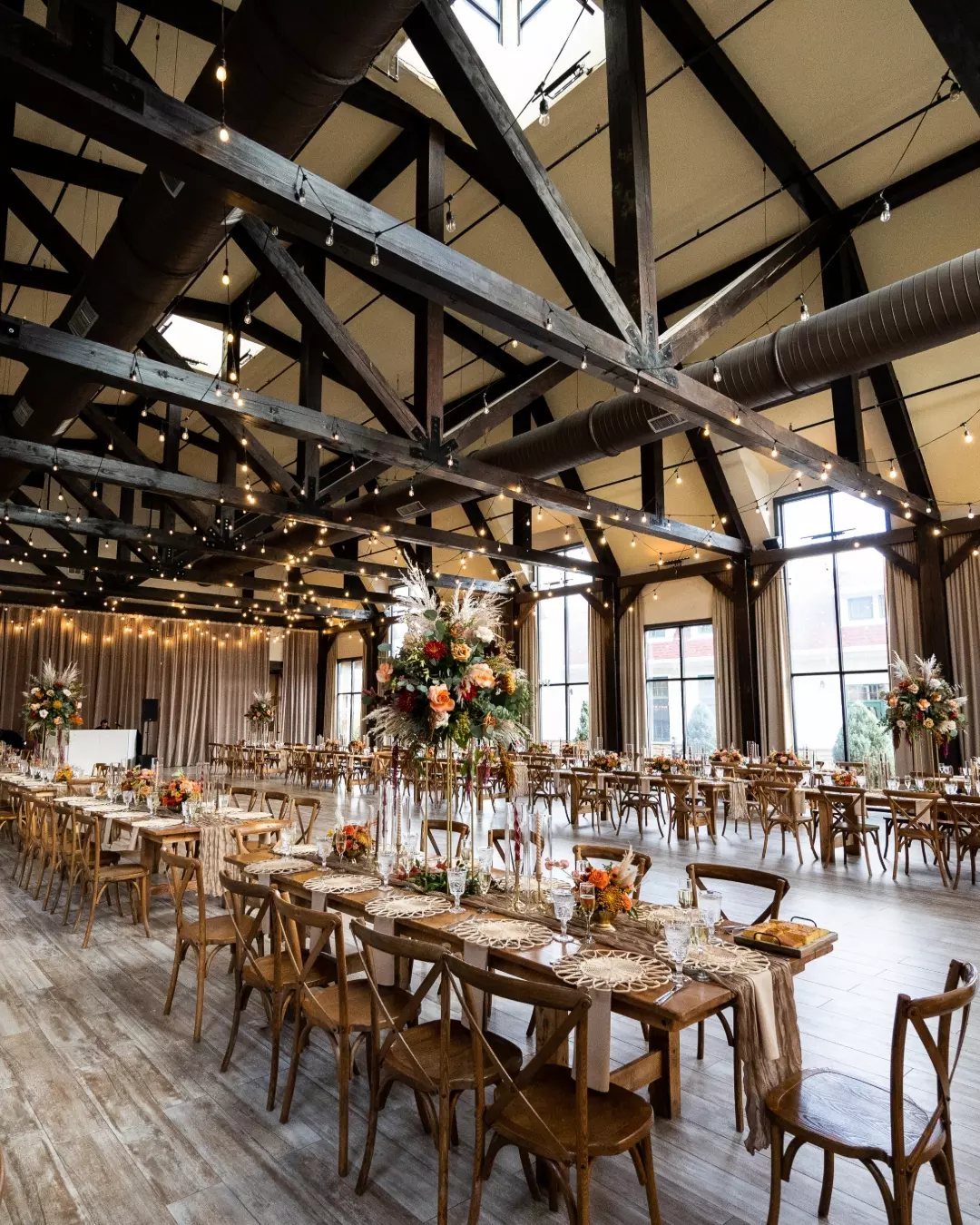 Wedding reception tables with floral centerpieces under dark wooden beams and string lights.
