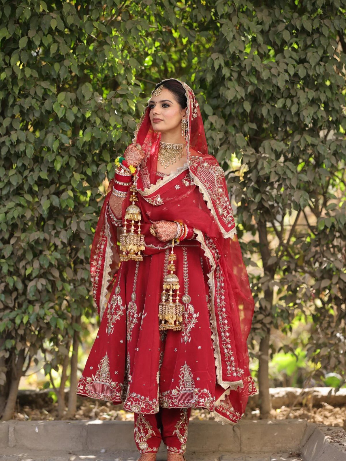 Woman in a red embroidered Anarkali suit and bridal jewelry against green foliage